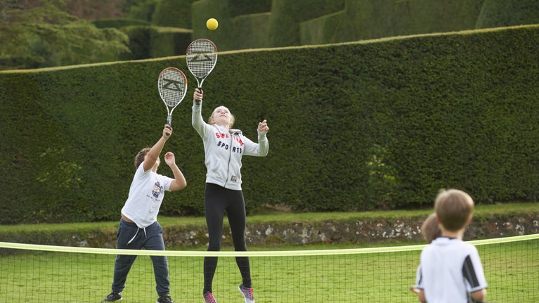Four children playing tennis at Chirk Castle, Wrexham. The two on the far side of the net stretch up with their racquets for the ball
